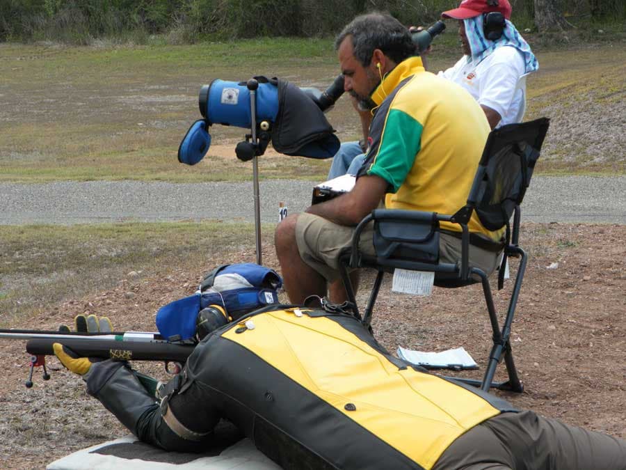 A shooter prone in a leather shooting jacket while a coach seated behind him reads the wind through a spotting scope