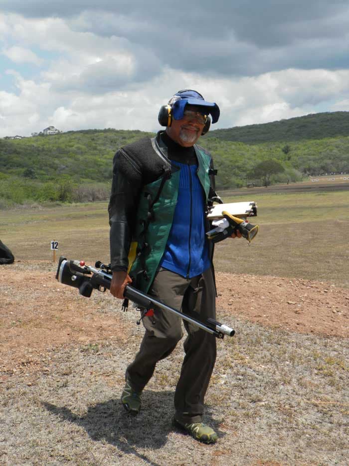 A shooter walking off the firing point in a shooting jacket and ear defenders, carrying his rifle and scoring board