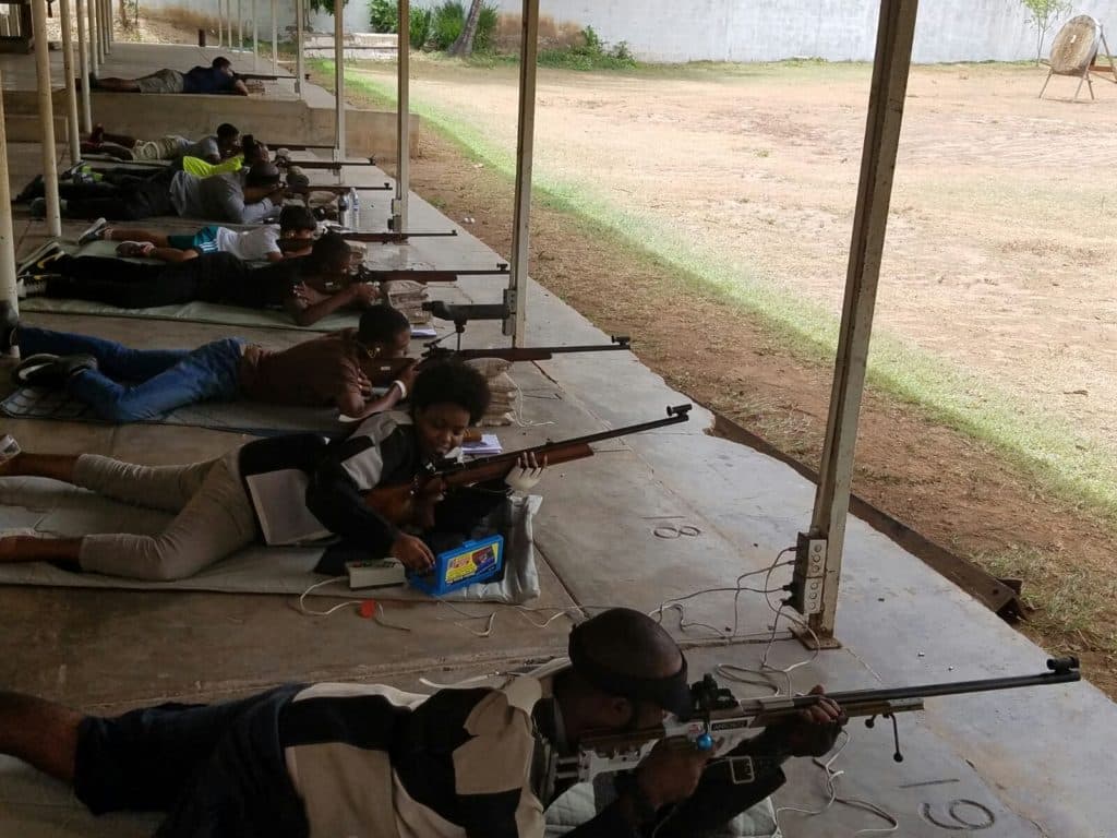 A squad of shooters lying prone in a row under the covered range, each with a smallbore rifle on the firing point