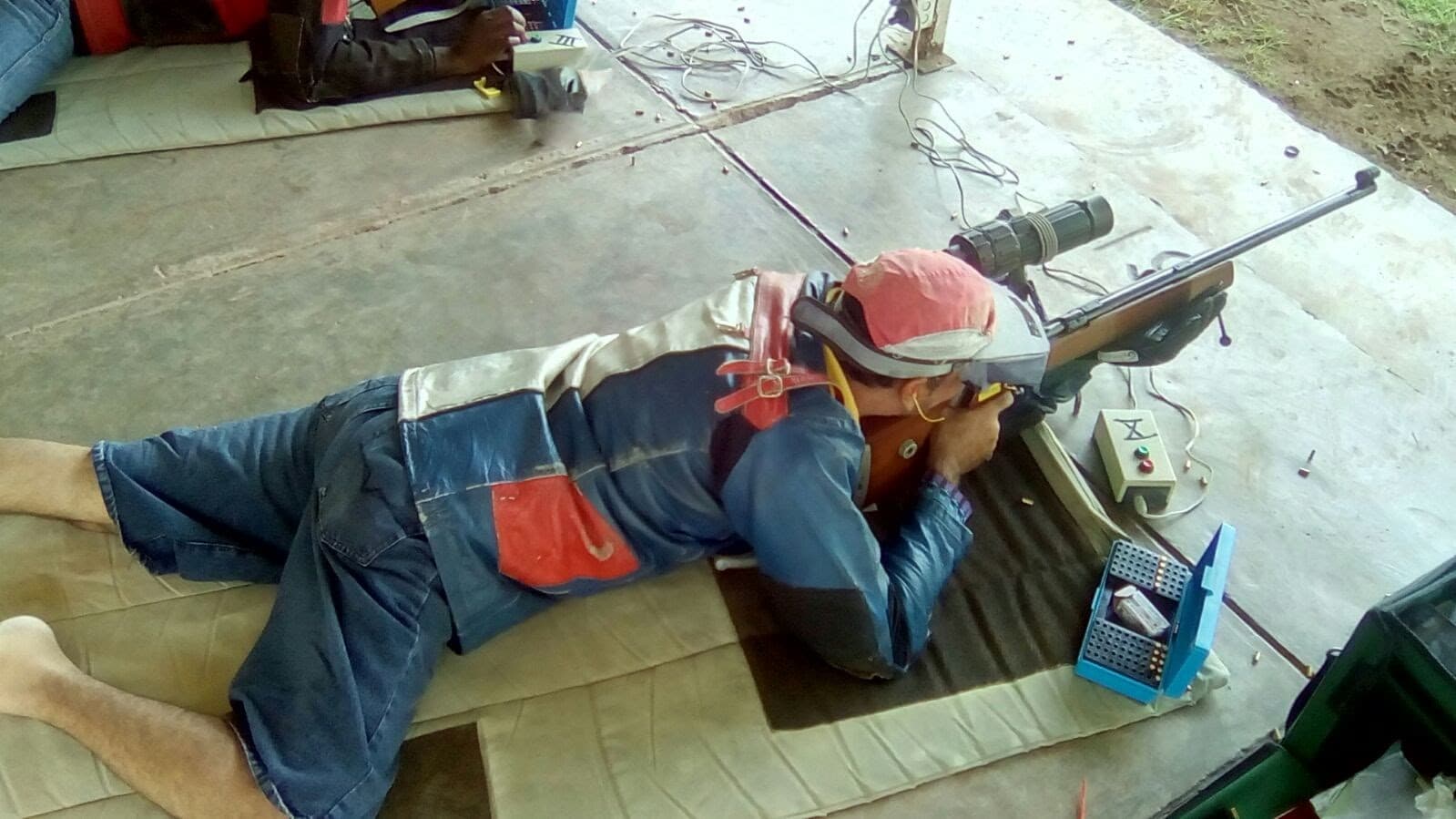 Overhead view of a shooter prone in a shooting jacket, an ammunition block and firing point controls beside the mat