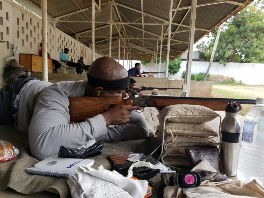 Close view of a shooter prone behind a smallbore rifle, sandbags, ammunition and a scoring pad laid out beside him