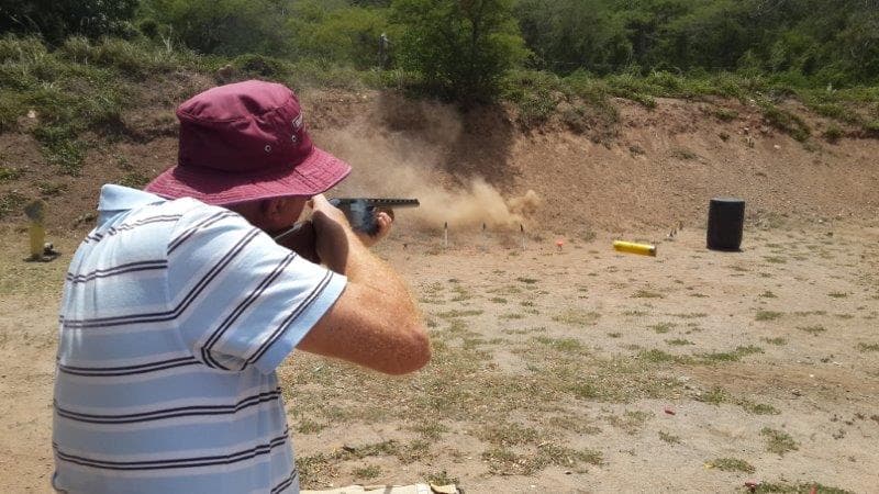 A shooter firing from the standing position at a steel target, dust kicking off the backstop behind it